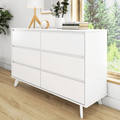 White modern six-drawer dresser with mid-century wooden legs on hardwood floor, minimalist bedroom storage staged with lamp, plant and books by sunny window