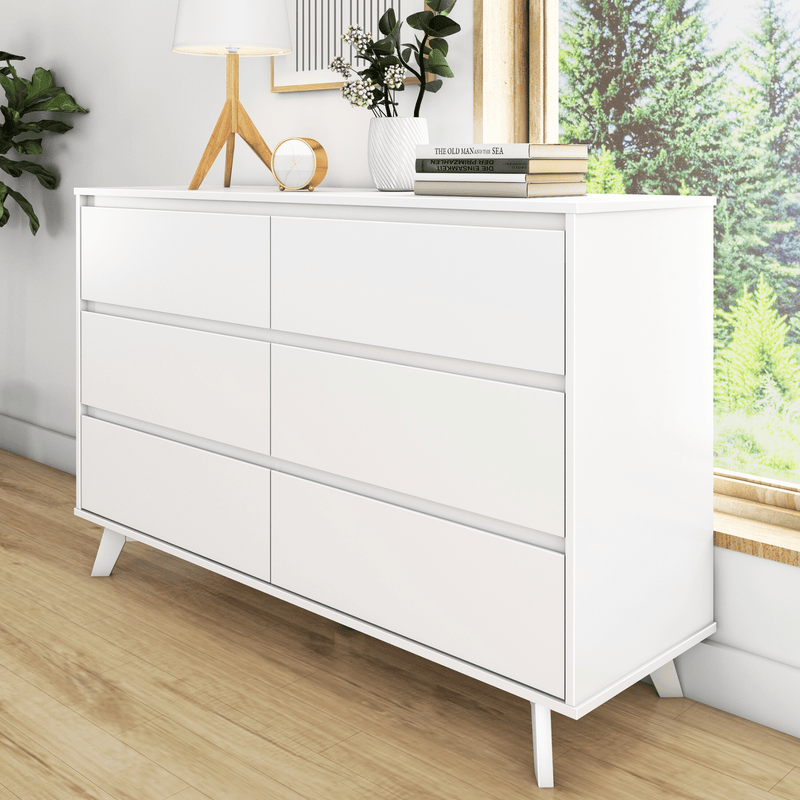 White modern six-drawer dresser with mid-century wooden legs on hardwood floor, minimalist bedroom storage staged with lamp, plant and books by sunny window