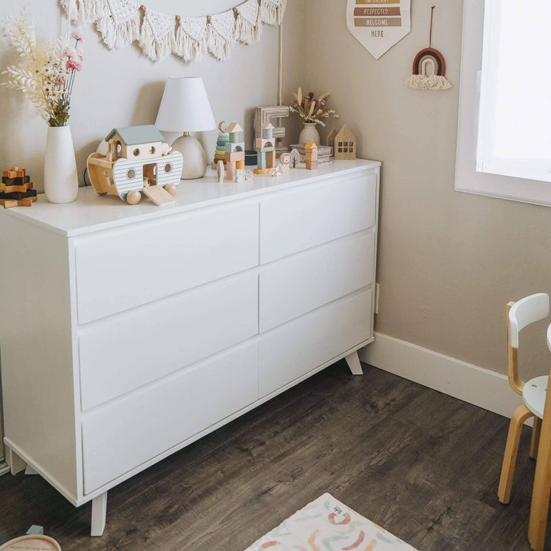 Modern white six-drawer dresser in a neutral boho nursery with wooden toys, table lamp, macramé wall hanging, small play chair and dark wood floor