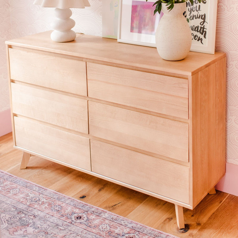 Light wood mid-century modern dresser with six drawers in pastel bedroom, decorative lamp and ceramic vase on top, hardwood floor and patterned rug