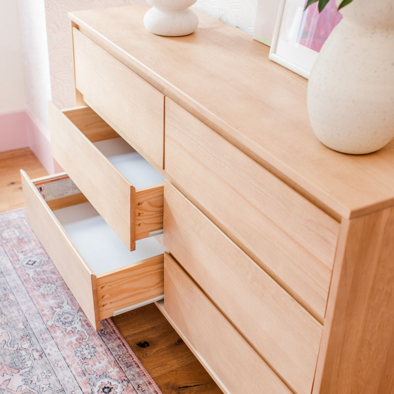 Wide light wood dresser with two open drawers, ceramic vases on top, pink patterned rug and hardwood floor in a minimalist bedroom.