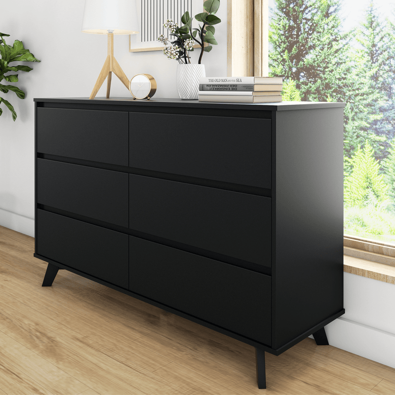 Modern matte black six-drawer dresser with angled legs and sleek handleless design for bedroom storage, styled with lamp and books by a sunlit window
