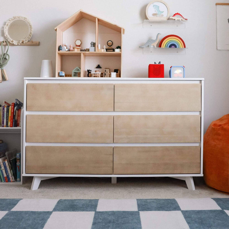 Modern white-framed wooden six-drawer nursery dresser with natural wood fronts, dollhouse and toys on top, rainbow wall shelf, kids books, checkered rug and orange beanbag