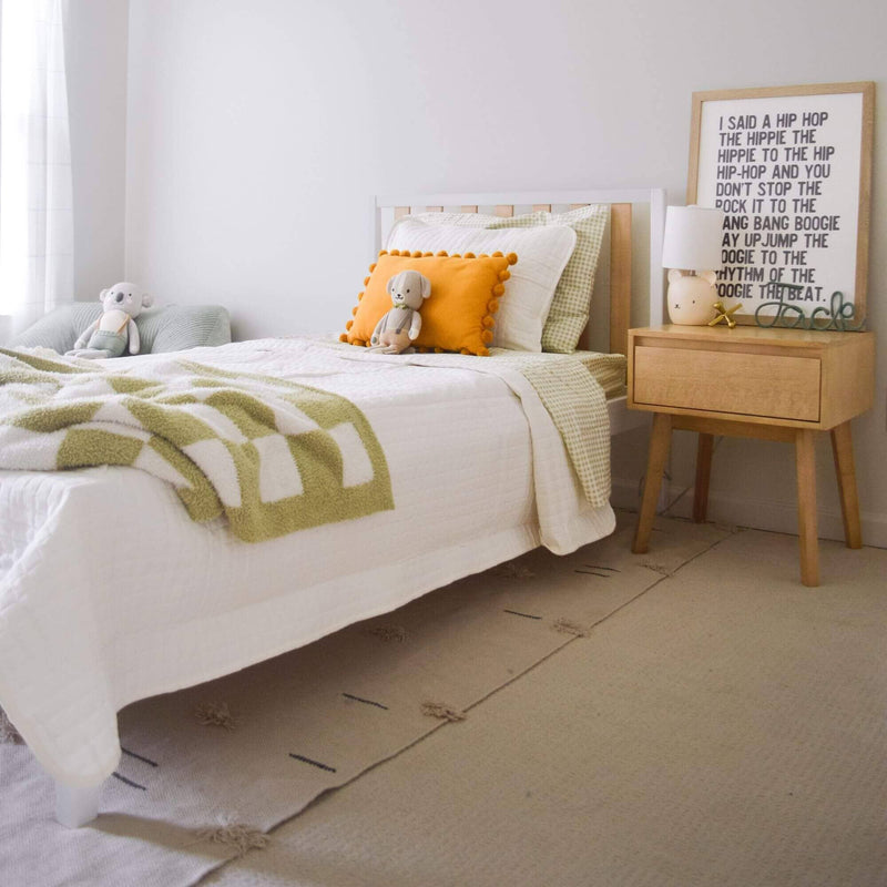 Cozy neutral kids bedroom with white toddler bed, green check bedding, orange pom-pom pillow, stuffed animals, wooden nightstand, lamp and framed typographic wall art