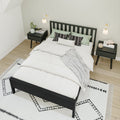 Modern minimalist bedroom with black slatted bed frame, white duvet, green and patterned accent pillows, matching black nightstands, wall sconces and geometric rug on light wood floor