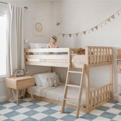 Light wooden bunk bed in modern neutral kids bedroom with smiling child on top bunk, checkered blue rug, bunting garland, cozy bedding and stuffed animals