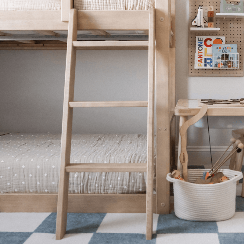 Natural wood bunk bed ladder in a neutral Scandinavian kids bedroom with quilted bedding, toy basket and pegboard book display.