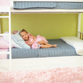 Toddler girl in pink dress lounging on a lower bunk bed with blue quilt in a pastel kids’ bedroom