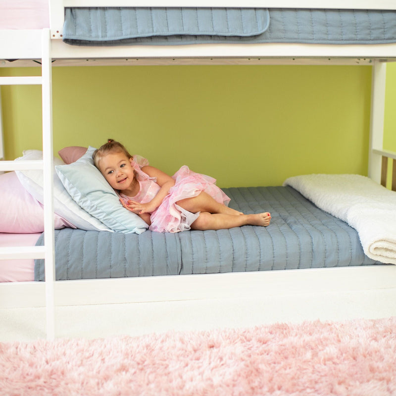 Toddler girl in pink dress lounging on a lower bunk bed with blue quilt in a pastel kids’ bedroom
