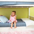 Smiling toddler girl in a pink dress sitting on a blue-quilted lower bunk bed in a green nursery with pillows and a pink shag rug — kids bedroom decor and bedding