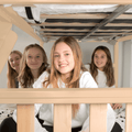 Four smiling girls posing under a natural wood bunk bed with white bedding in a cozy bedroom sleepover scene