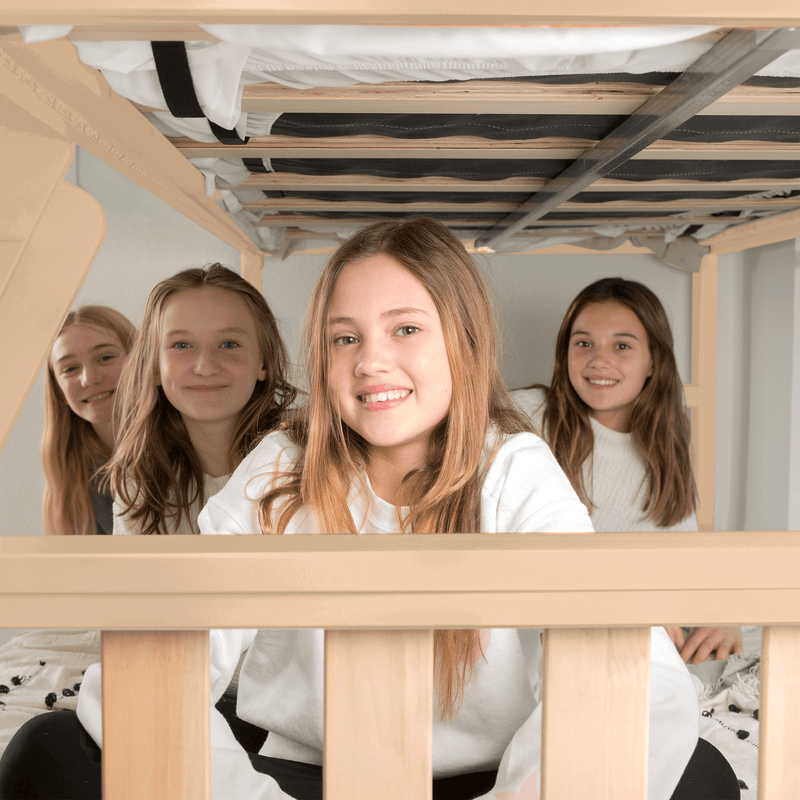 Four smiling girls posing under a natural wood bunk bed with white bedding in a cozy bedroom sleepover scene