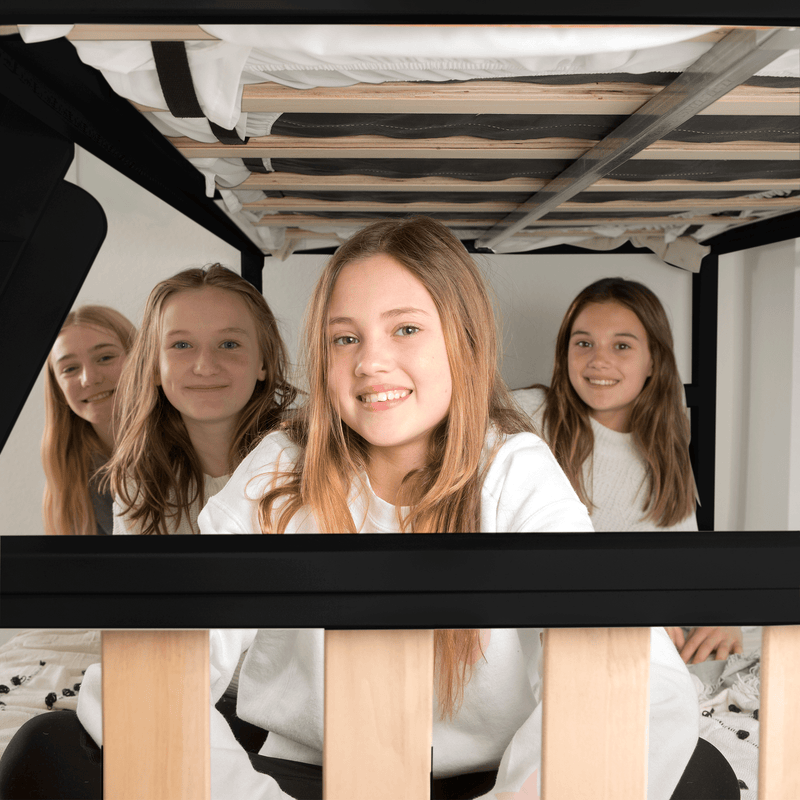Smiling girls huddled under a bunk bed at a sleepover, close-up faces framed by wooden bed slats and mattress supports