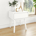 White modern bedside table nightstand with single drawer and wooden legs, lamp, vase with greenery and gold alarm clock on hardwood floor by window with forest view