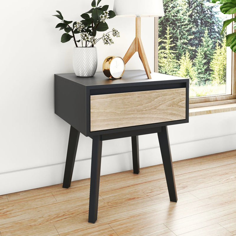 Mid-century modern black and light wood nightstand with single drawer and tapered legs, styled with lamp, clock, and plant by a window