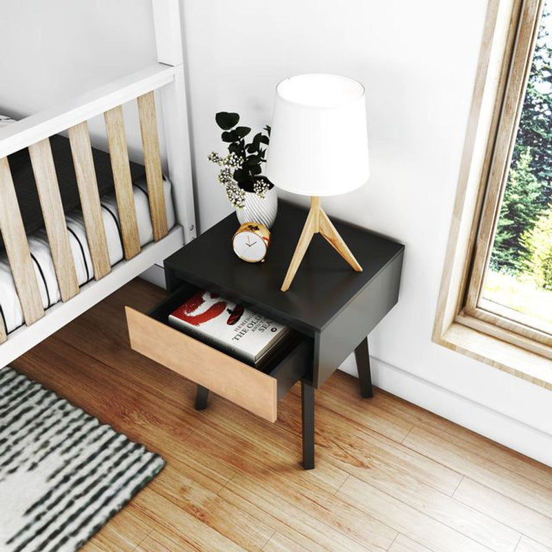 Modern black-and-wood bedside table nightstand with open wooden drawer, tripod table lamp, alarm clock and plant vase next to a crib on hardwood floor by a sunlit window