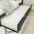 Pull-out trundle bed under a modern black wooden frame with white bedding and gray fringed throw on hardwood floor with patterned rug