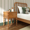 Wooden nightstand with turned spindle legs beside a spindle headboard bed in a children's bedroom, white bedding, green throw pillow, teddy bear, toy camera and pencil holder on top, striped wallpaper and hardwood floor.