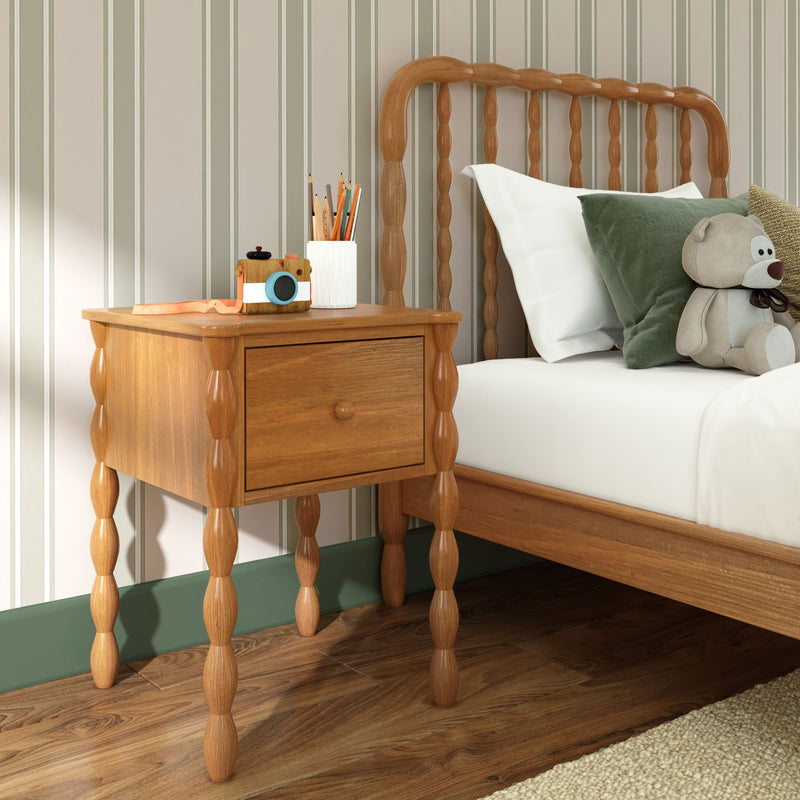 Wooden nightstand with turned spindle legs beside a spindle headboard bed in a children's bedroom, white bedding, green throw pillow, teddy bear, toy camera and pencil holder on top, striped wallpaper and hardwood floor.