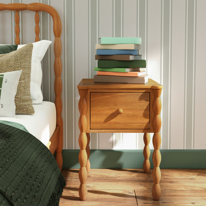 Wooden turned-leg bedside nightstand with stacked books next to bed with green quilt and textured pillows in a cozy farmhouse bedroom