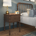 Dark wooden turned-leg bedside table with brass table lamp, wooden toy camera, notebook and plush frog pillow beside a spindle headboard on a hardwood floor with patterned rug