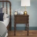 Wooden spindle-leg nightstand with brass table lamp, vintage alarm clock and plush whale beside a cozy bed in blue bedroom