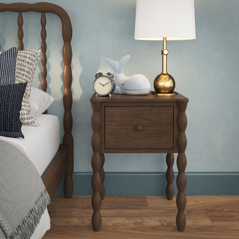 Wooden spindle-leg nightstand with brass table lamp, vintage alarm clock and plush whale beside a cozy bed in blue bedroom