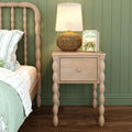 Cozy cottage-style bedroom nightstand with light wood spindle bedside table, woven rattan lamp, and gardening books beside a matching spindle bed against a sage green paneled wall