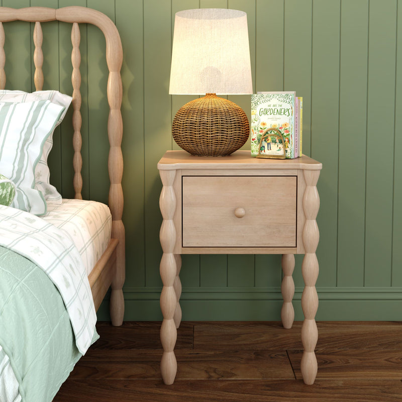 Cozy cottage-style bedroom nightstand with light wood spindle bedside table, woven rattan lamp, and gardening books beside a matching spindle bed against a sage green paneled wall