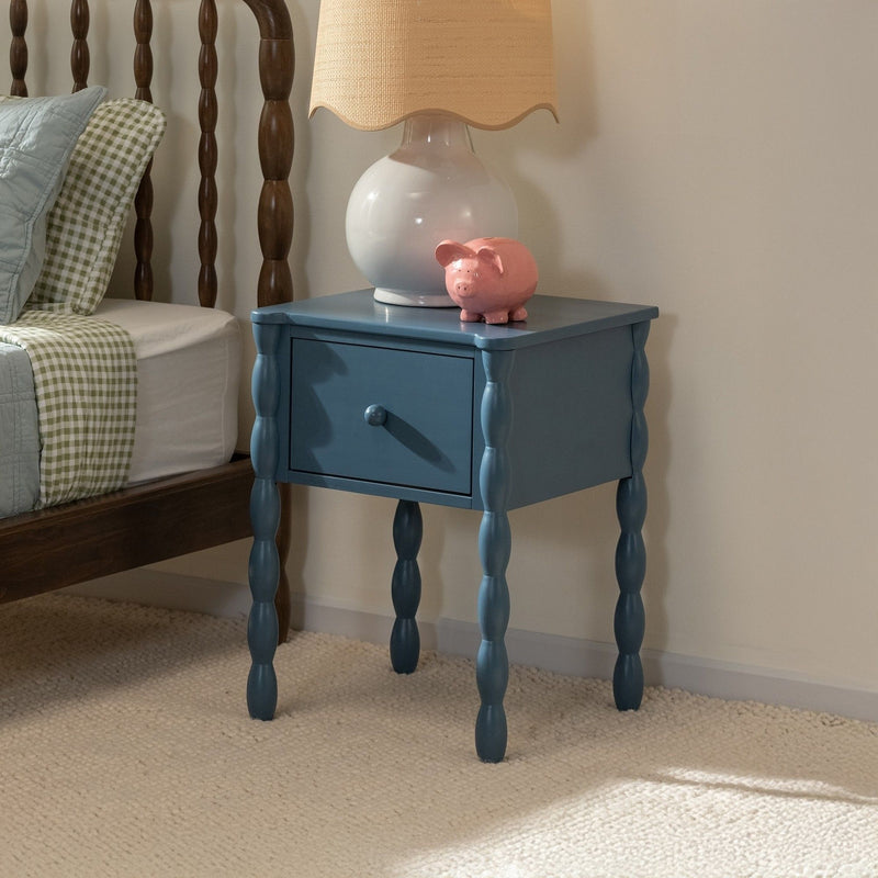 Blue turned-leg bedside table with drawer, white ceramic lamp and pink piggy bank beside wooden spindle bed with green gingham bedding in cozy bedroom