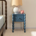 Blue wooden bedside table nightstand with turned legs and drawer, white ceramic lamp with beige shade and pink piggy bank beside wooden spindle bed in cozy bedroom