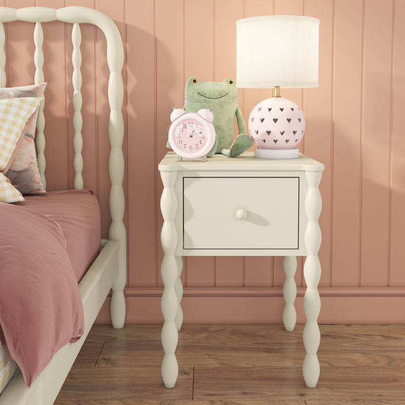 Cream spindle nightstand in pastel pink kids' bedroom with heart-cut pink table lamp, pink alarm clock, green frog plush, white spindle bed and wood floor