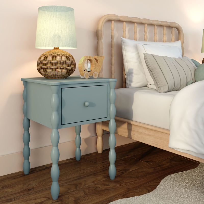 Blue spindle-leg bedside table with drawer, rattan lamp and wooden toy elephant beside a light wood spindle bed with neutral pillows and hardwood floor