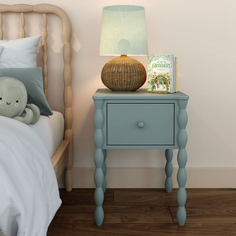 Blue bedside table with drawer and spindle legs topped by a woven rattan lamp, children's books and plush octopus beside a wooden bed — kids bedroom nursery decor