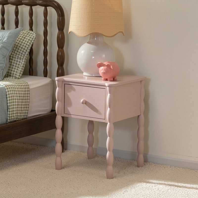Pink bedside table nightstand with turned spindle legs and single drawer, topped by a white ceramic lamp and pink piggy bank beside a wooden spindle bed with green gingham bedding on a carpeted floor