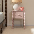 Pink nightstand bedside table with turned spindle legs and single drawer topped with a white ceramic lamp and pink piggy bank, cozy bedroom decor