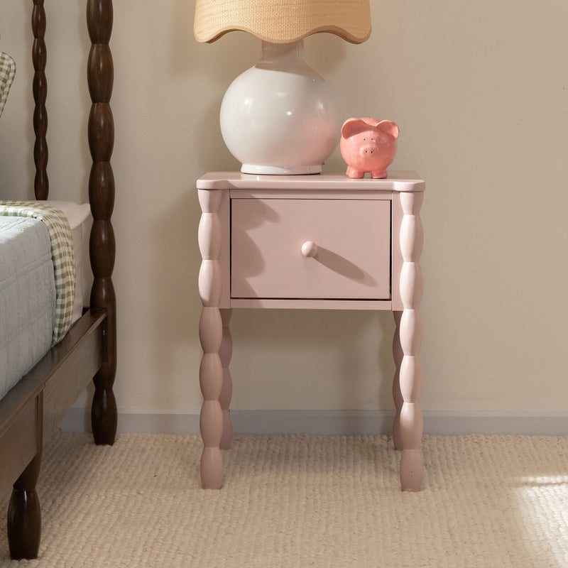 Pink nightstand bedside table with turned spindle legs and single drawer topped with a white ceramic lamp and pink piggy bank, cozy bedroom decor