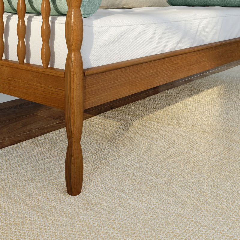 Close-up of solid wood spindle bed frame and white mattress on hardwood floor with beige rug and green bolster pillow visible