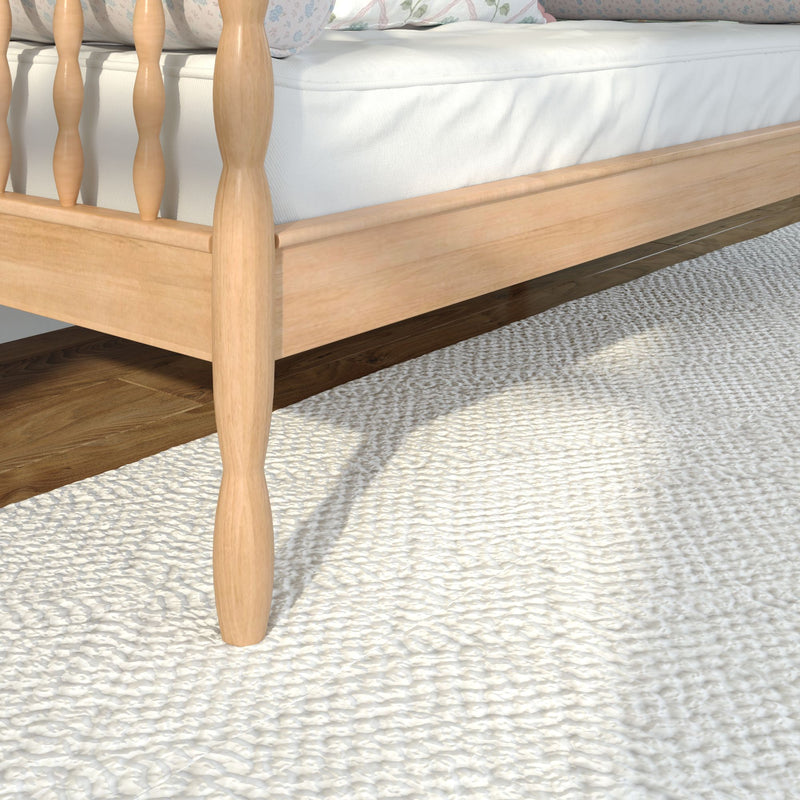 Close-up of light wood spindle bed frame with white mattress on a textured cream rug — Scandinavian-style bedroom furniture and cozy bedding detail