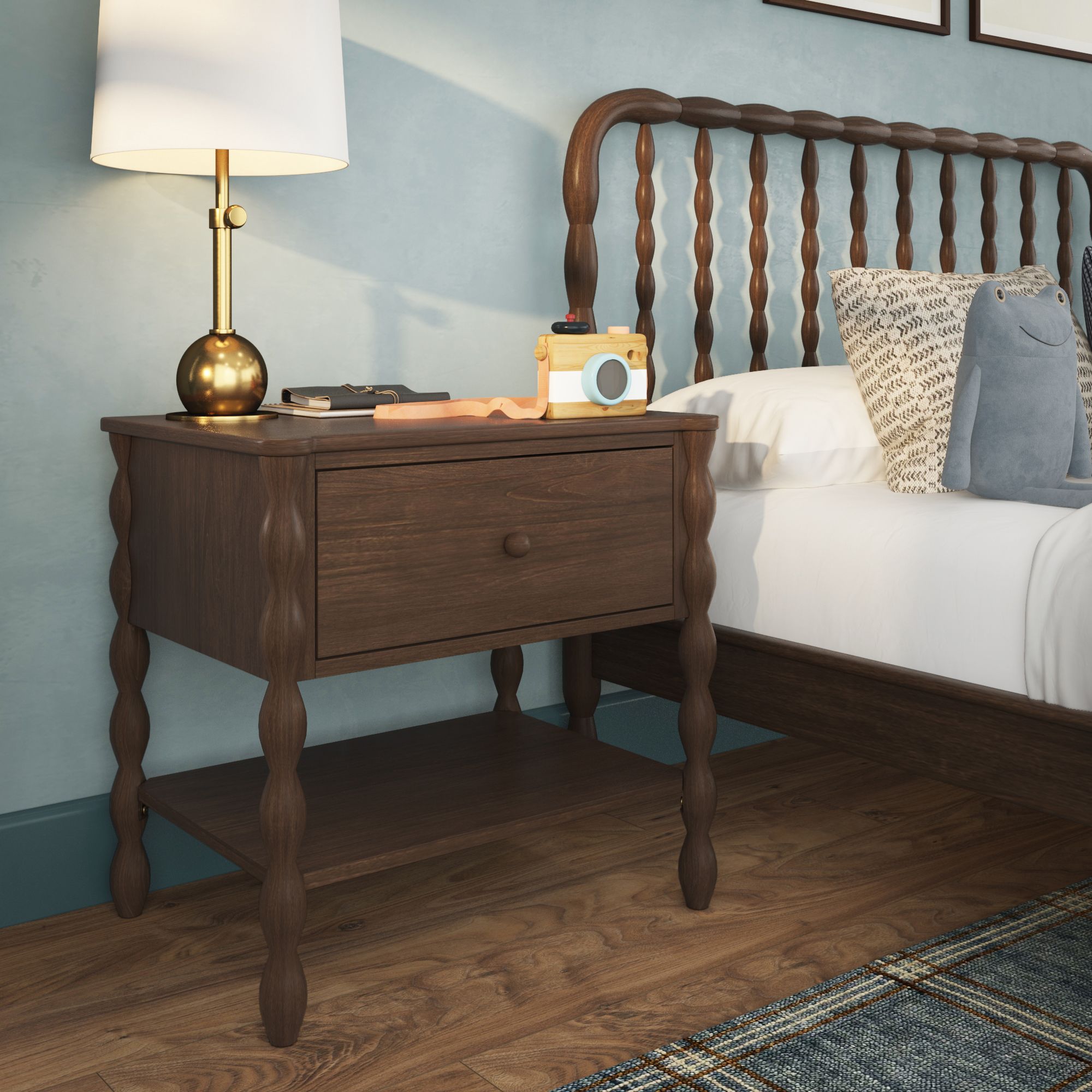 A dark-wood finish Jenny Lind style nightstand with one drawer and shelf next to a matching bed. There is a lamp and decor on the nightstand.