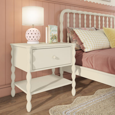 White spindle nightstand with drawer and lower shelf beside matching white spindle bed in a pink paneled girls' bedroom, heart-cutout ceramic lamp, gingham pillow, floral book, wood floor and braided jute rug