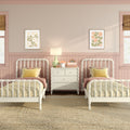 Pastel pink twin girls' bedroom with white spindle beds, yellow pillows, gingham bedding, scalloped wainscoting, central nightstand with lamp and framed floral wall art