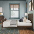 Coastal bedroom interior with wooden spindle bed and tufted headboard, blue-gray walls, window framing a beach view, nautical decor, plaid area rug and bedside lamp