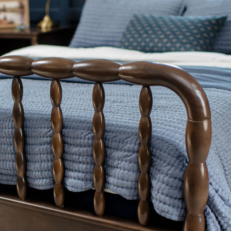 Close-up of dark wooden spindle bed frame with blue quilted bedding and patterned accent pillow, cozy bedroom decor