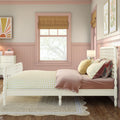 Cozy pastel pink girls' bedroom with white spindle bed, yellow gingham bedding, layered decorative pillows, scalloped wainscoting, roman shade and framed botanical art