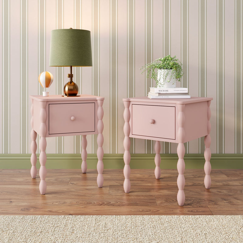 Pastel pink bedside tables with turned spindle legs against beige-and-sage striped wallpaper, styled with a green fabric table lamp, small hot-air-balloon ornament and a potted trailing plant on stacked macrame books — cozy vintage-inspired bedroom decor