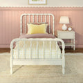 Pastel pink children's bedroom with white spindle bed, yellow gingham bedding and pillow, scalloped wainscoting and bedside table with lamp and alarm clock