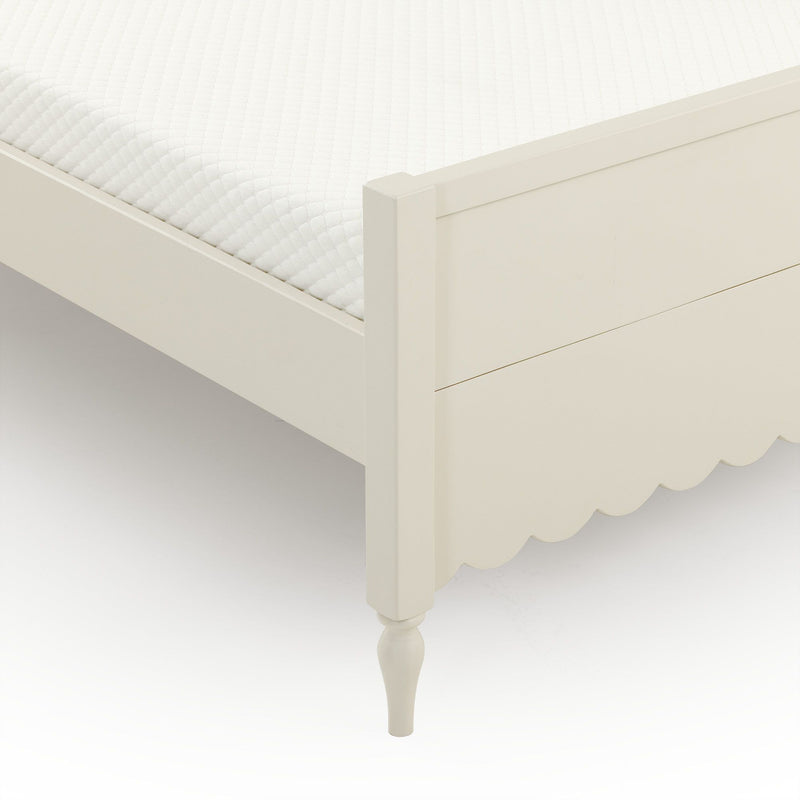 Close-up of white quilted mattress topper on cream wooden bed frame with scalloped skirt and turned leg, minimalist bedroom furniture