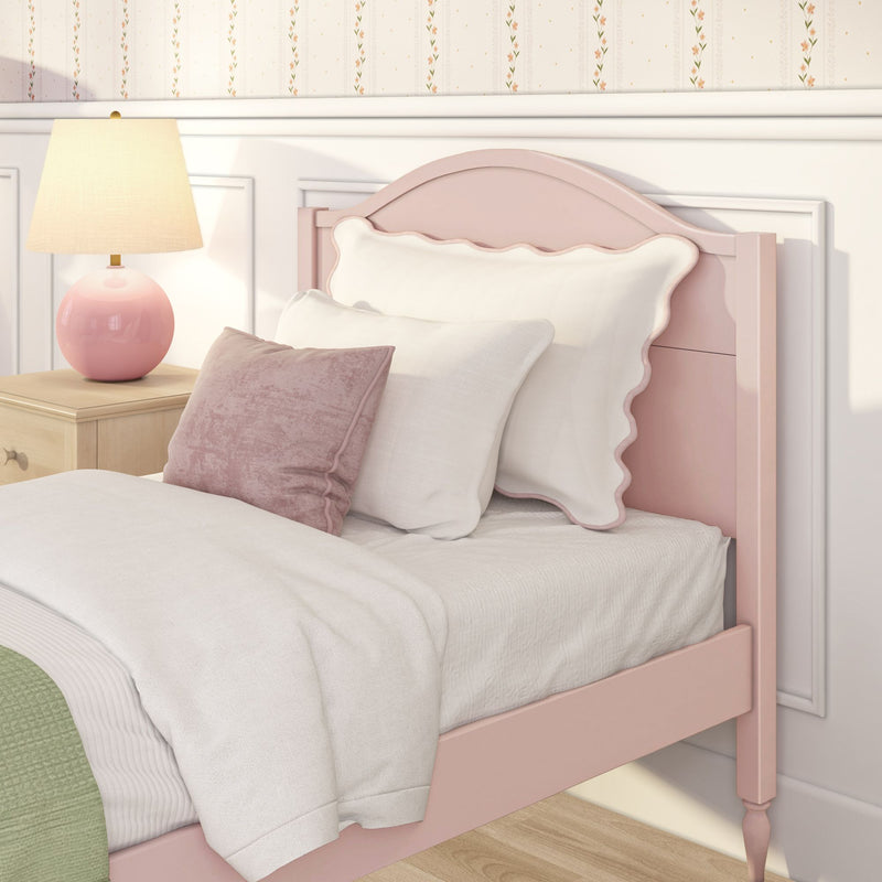 Pastel pink girls' bedroom with scalloped pink headboard, white linens and blush pillows, pink ceramic bedside lamp on a wooden nightstand and floral wallpaper — cozy vintage nursery decor