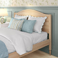 Cozy coastal bedroom close-up of natural wood bed frame with white scalloped pillows blue throw pillows floral wallpaper pastel wainscoting and bedside lamp